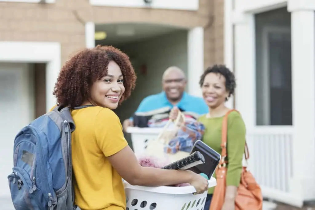Mature African-American parents helping their daughter relocate, perhaps into an apartment or college dorm. The young woman is in the foreground smiling at the camera, carrying a backpack and basket filled with her belongings.