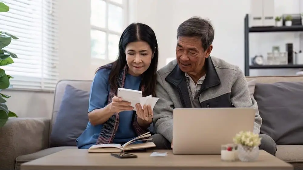 Asian senior couple checking and calculate financial bill together on sofa involved in financial paperwork, paying tax online using laptop at living room home